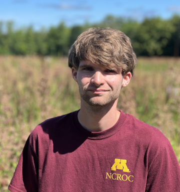 A man in a university of minnesota shirt stands in front of wild rice.
