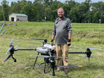 A man standing in front of a large drone with fields and a shed in the background.